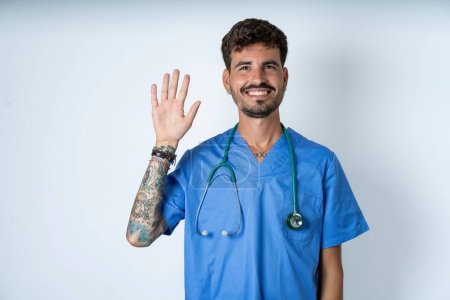 Foto de Guapo enfermero vistiendo uniforme de cirujano sobre fondo blanco mostrando y apuntando hacia arriba con los dedos número cinco mientras sonríe confiado y feliz. - Imagen libre de derechos