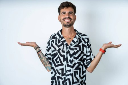 Téléchargez les photos : Joyeux homme optimiste joyeux avec tatouage portant chemise d'été debout sur fond blanc isolé tenant deux paumes espace de copie - en image libre de droit