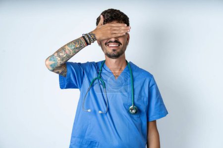 Foto de Guapo enfermero vistiendo uniforme de cirujano sobre fondo blanco sonriendo y riendo con la mano en la cara cubriendo los ojos para sorpresa. Concepto ciego. - Imagen libre de derechos