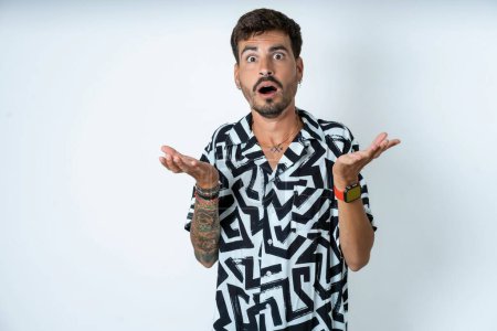Photo for Frustrated man with tattoo wearing summer shirt standing over isolated white background feels puzzled and hesitant, shrugs shoulders in bewilderment, keeps mouth widely opened, doesn't know what to do. - Royalty Free Image