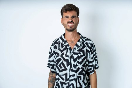 Photo for Man with tattoo wearing summer shirt standing over isolated white background keeps teeth clenched, frowns face in dissatisfaction, irritated because of much duties. - Royalty Free Image