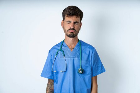 Photo for Dismal gloomy rejected handsome nurse man wearing surgeon uniform over white background has problems and difficulties, curves lower lip and closes eyes in despair, being in depression - Royalty Free Image