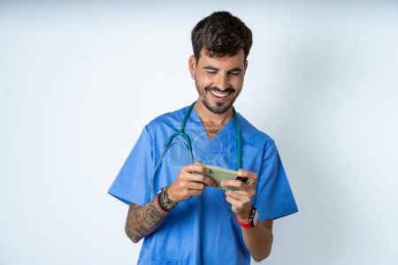 Photo for Handsome nurse man wearing surgeon uniform over white background holding in hands cell playing video games or chatting - Royalty Free Image