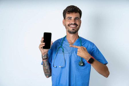 Foto de Atractivo alegre guapo enfermero vistiendo uniforme cirujano sobre fondo blanco sosteniendo en las manos celda mostrando pantalla negra - Imagen libre de derechos