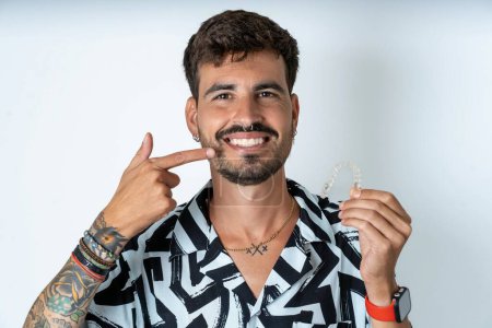 Téléchargez les photos : Jeune homme caucasien portant une chemise imprimée tenant un aligneur invisible et pointant des dents droites parfaites. Concept de soins dentaires et de confiance. - en image libre de droit