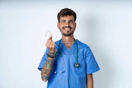 Photo for Handsome nurse man wearing surgeon uniform over white background holding an invisible braces aligner, recommending this new treatment. Dental healthcare concept. - Royalty Free Image