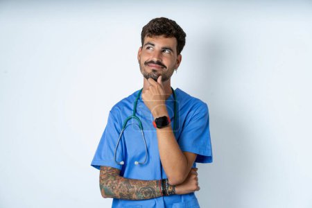 Foto de Pensativo guapo enfermero vistiendo uniforme de cirujano sobre fondo blanco sostiene la barbilla y mira hacia otro lado pensativamente hace un gran plan - Imagen libre de derechos