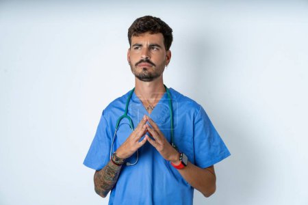 Foto de Guapo enfermero vistiendo uniforme de cirujano sobre fondo blanco dedos inclinados y se ve misterioso a un lado tiene gran plan malvado en mente - Imagen libre de derechos