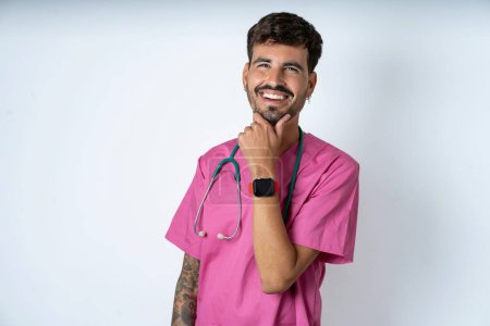Foto de Guapo enfermero vistiendo uniforme de cirujano sobre fondo blanco risas felizmente mantiene la mano en la barbilla expresa emociones positivas sonrisas ampliamente tiene expresión despreocupada - Imagen libre de derechos