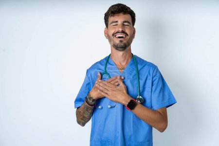 Foto de Guapo enfermero vistiendo uniforme de cirujano sobre fondo blanco expresa felicidad, ríe agradablemente, mantiene las manos en el corazón - Imagen libre de derechos