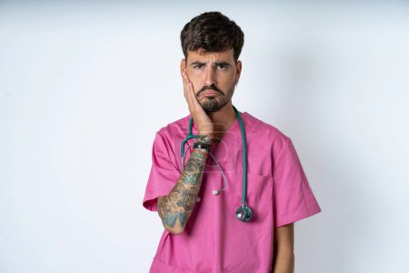 Photo for Sad lonely handsome nurse man wearing surgeon uniform over white background touches cheek with hand bites lower lip and gazes with displeasure. Bad emotions - Royalty Free Image