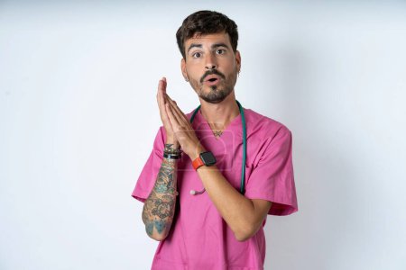 Téléchargez les photos : Surpris émotionnel beau infirmier homme portant un uniforme de chirurgien sur fond blanc frotte les paumes et regarde la caméra avec incrédulité - en image libre de droit