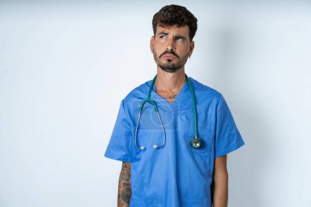 Photo for Dissatisfied handsome nurse man wearing surgeon uniform over white background purses lips and has unhappy expression looks away stands offended. Depressed frustrated model. - Royalty Free Image