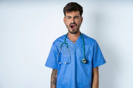 Foto de El hombre guapo de la enfermera que usa uniforme del cirujano sobre bostezos blancos del fondo con los puestos abiertos de la boca. Rutina diaria - Imagen libre de derechos
