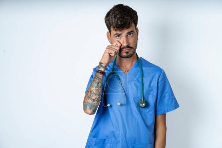 Photo for Disappointed dejected handsome nurse man wearing surgeon uniform over white background wipes tears stands stressed with gloomy expression. Negative emotion - Royalty Free Image