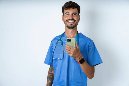 Foto de Guapo enfermero vistiendo uniforme cirujano sobre fondo blanco sostiene el teléfono móvil en las manos y se alegra de noticias positivas, utiliza celular moderno - Imagen libre de derechos