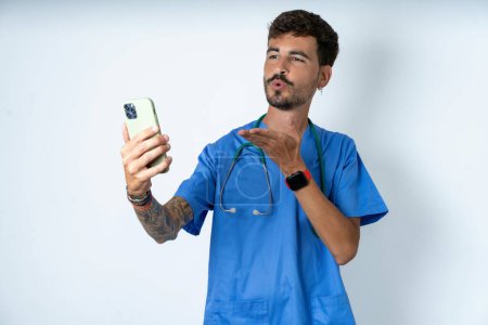 Téléchargez les photos : Belle infirmière homme portant uniforme de chirurgien sur fond blanc souffle baiser d'air à la caméra du smartphone et prend selfie, envoie mwah via appel en ligne. - en image libre de droit