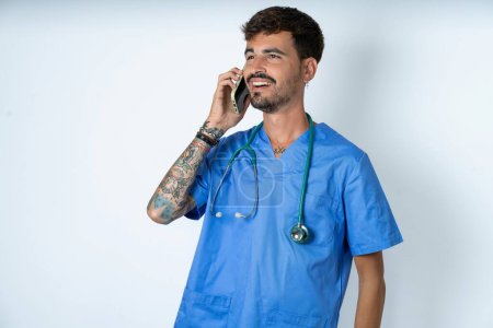 Foto de Agradable mirada feliz guapo enfermero vistiendo uniforme cirujano sobre fondo blanco tiene buena conversación telefónica y mira a un lado, tiene buen humor y sonríe positivamente mientras habla a través del teléfono celular - Imagen libre de derechos