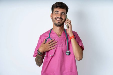 Foto de Sonriente enfermero guapo vistiendo uniforme de cirujano sobre fondo blanco habla a través del teléfono celular, disfruta de una agradable gran conversación. Personas, tecnología, concepto de comunicación - Imagen libre de derechos