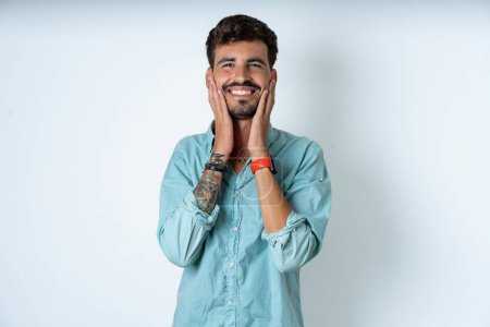 Photo for Happy handsome young man wearing turquoise shirt over white background touches both cheeks gently, has tender smile, shows white teeth, gazes positively straightly at camera, - Royalty Free Image