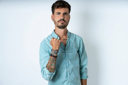 Photo for Handsome young man wearing turquoise shirt over white background shows fist has annoyed face expression going to revenge or threaten someone makes serious look. I will show you who is boss - Royalty Free Image