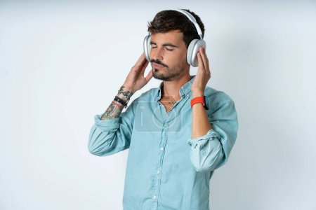 Photo for Handsome young man wearing turquoise shirt over white background with headphones on her head, listens to music, enjoying favourite song with closed eyes, holding hands on headset. - Royalty Free Image