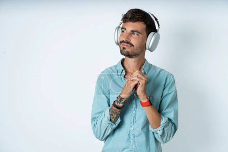 Photo for Handsome young man wearing turquoise shirt over white background wears stereo headphones listening to music concentrated and looking aside with interest. - Royalty Free Image