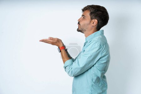 Téléchargez les photos : Profil vue de côté, portrait d'un beau jeune homme attrayant portant une chemise turquoise sur fond blanc envoyant un baiser d'air - en image libre de droit
