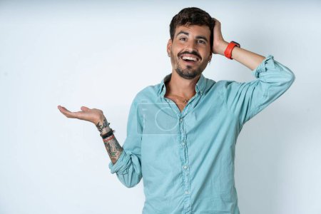 Foto de Sorprendido sorprendido sorprendido joven guapo con camisa de color turquesa sobre fondo blanco sostener la mano ofreciendo proposición - Imagen libre de derechos