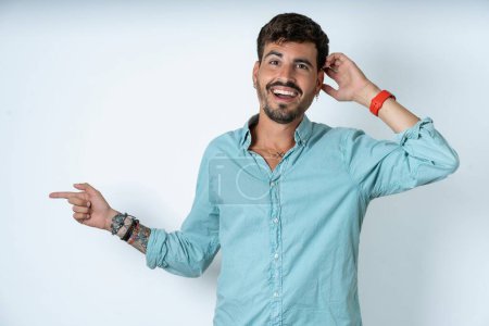 Photo for Surprised handsome young man wearing turquoise shirt over white background pointing at empty space holding hand on head - Royalty Free Image