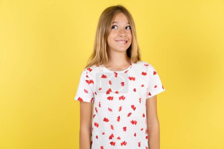 Téléchargez les photos : Incroyable enfant caucasien fille portant chemise à pois sur fond jaune lèvre mordante et regardant délicat à vide espace. - en image libre de droit