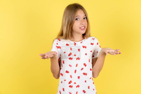 Photo for Clueless caucasian kid girl wearing polka dot shirt over yellow background shrugs shoulders with hesitation, faces doubtful situation, spreads palms, Hard decision - Royalty Free Image