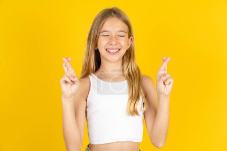 Photo for Blonde girl wearing white T-shirt over yellow background has big hope, crosses fingers, believes in good fortune, smiles broadly. People and wish concept - Royalty Free Image