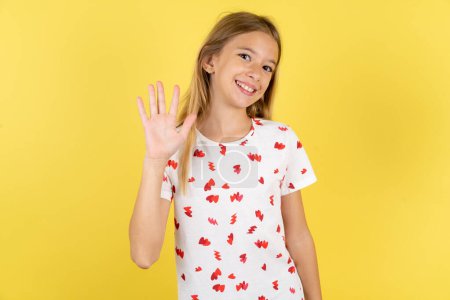 Foto de Niña caucásica con camisa de lunares sobre fondo amarillo Renuncia a decir hola feliz y sonriente, gesto de bienvenida amistoso. - Imagen libre de derechos
