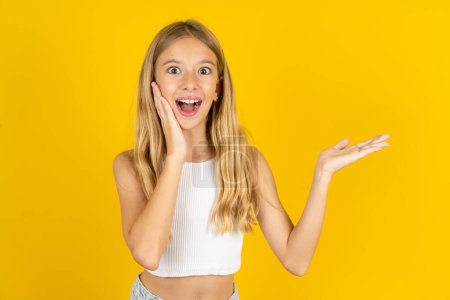 Crazy blonde girl wearing white T-shirt over yellow background advising discount prices hold open palm new product