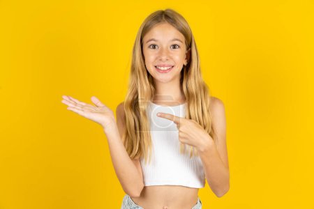 Téléchargez les photos : Blonde fille portant blanc T-shirt sur fond jaune pointant et tenant la main montrant annonces - en image libre de droit