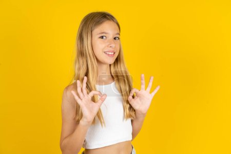 Foto de Chica rubia vistiendo camiseta blanca sobre fondo amarillo mostrando ambas manos con los dedos en signo OK. Aprobación o recomendación del concepto - Imagen libre de derechos
