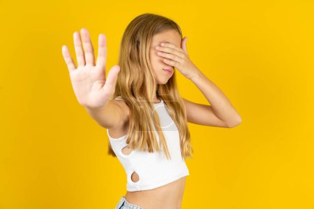 Photo for Blonde girl wearing white T-shirt over yellow background covers eyes with palm and doing stop gesture, tries to hide from everybody. - Royalty Free Image