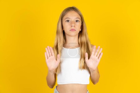 Photo for Serious blonde girl wearing white T-shirt over yellow background pulls palms towards camera, makes stop gesture, asks to control your emotions and not be nervous - Royalty Free Image