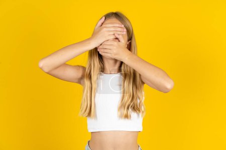 Photo for Blonde girl wearing white T-shirt over yellow background Covering eyes and mouth with hands, surprised and shocked. Hiding emotions. - Royalty Free Image