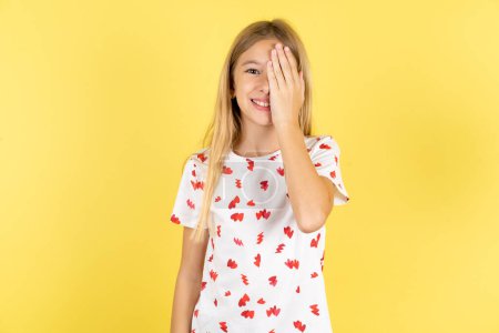 Téléchargez les photos : Enfant caucasien fille portant chemise à pois sur fond jaune couvrant un oeil avec sa main, sourire confiant sur le visage et l'émotion surprise. - en image libre de droit