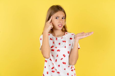 Photo for Caucasian kid girl wearing polka dot shirt over yellow background confused and annoyed with open palm showing copy space and pointing finger to forehead. Think about it. - Royalty Free Image