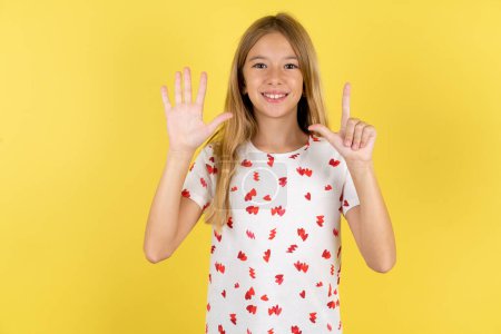 Foto de Niña caucásica con camisa de lunares sobre fondo amarillo mostrando y apuntando hacia arriba con los dedos número siete mientras sonríe confiado y feliz. - Imagen libre de derechos