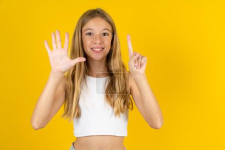 Foto de Chica rubia vistiendo camiseta blanca sobre fondo amarillo mostrando y apuntando hacia arriba con los dedos número seis mientras sonríe confiado y feliz. - Imagen libre de derechos