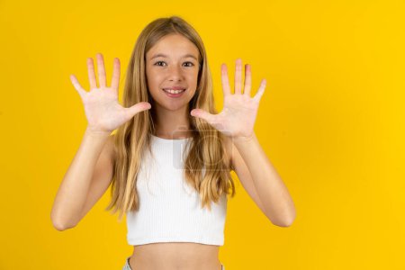 Foto de Chica rubia vistiendo camiseta blanca sobre fondo amarillo mostrando y apuntando hacia arriba con los dedos número diez mientras sonríe confiado y feliz. - Imagen libre de derechos