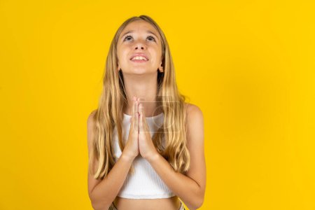 Téléchargez les photos : Fille blonde portant blanc T-shirt sur fond jaune mendier et prier avec les mains ainsi que l'expression d'espoir sur le visage très émotionnel et inquiet. Mon Dieu ! - en image libre de droit