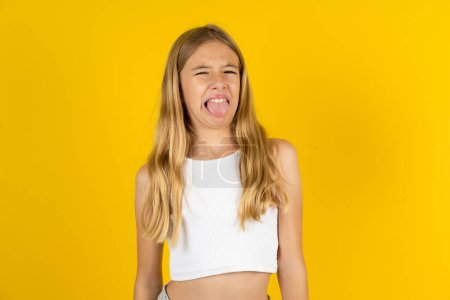 Blondes Mädchen in weißem T-Shirt über gelbem Hintergrund streckt die Zunge glücklich mit lustigem Gesichtsausdruck heraus. Emotionales Konzept.