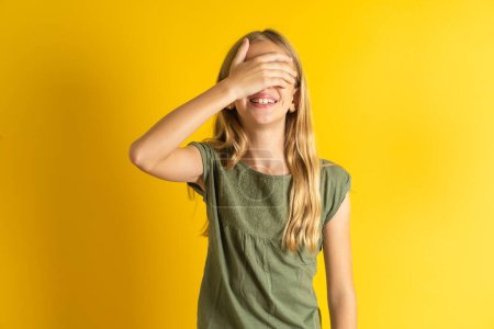 Photo for Blond little girl wearing khaki blouse over yellow background smiling and laughing with hand on face covering eyes for surprise. Blind concept. - Royalty Free Image