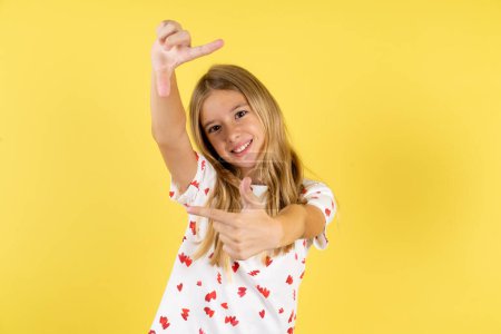 Téléchargez les photos : Fille enfant caucasien portant chemise à pois sur fond jaune faisant cadre doigt avec les mains. Concept de créativité et de photographie. - en image libre de droit