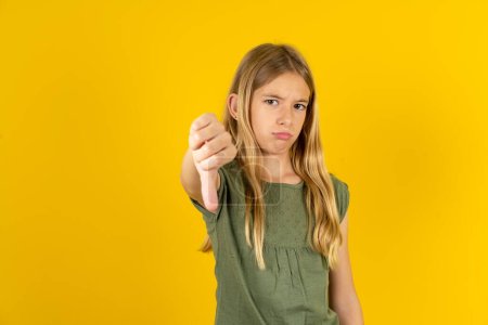 Téléchargez les photos : Blond petite fille portant chemisier kaki sur fond jaune se sentant en colère, ennuyé, déçu ou mécontent, montrant pouces vers le bas avec un regard sérieux - en image libre de droit